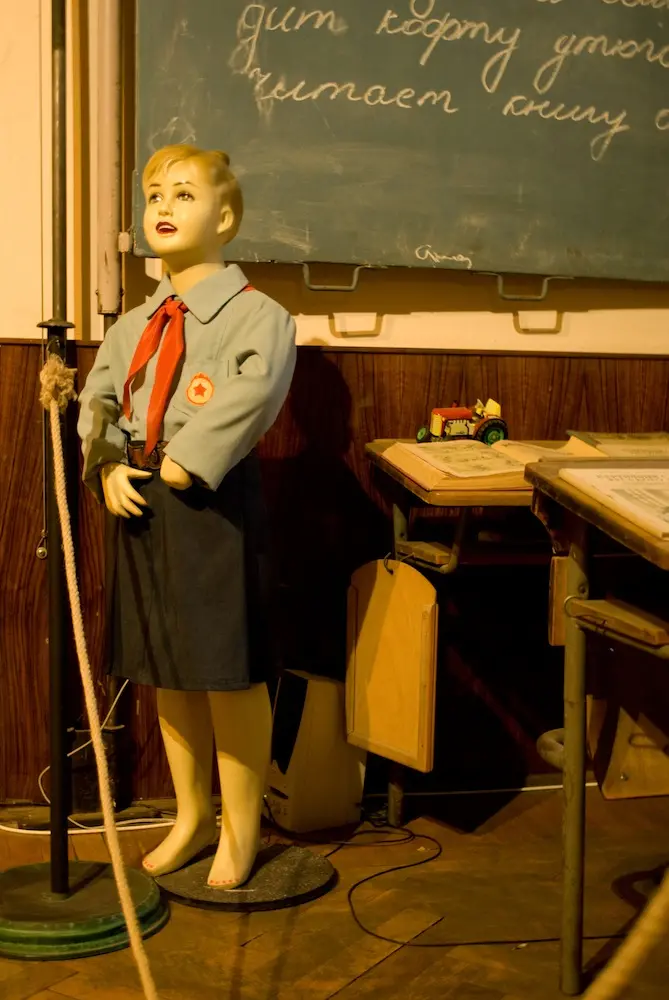 A plastic doll dressed up in Soviet classroom attire standing in front of a chalk board