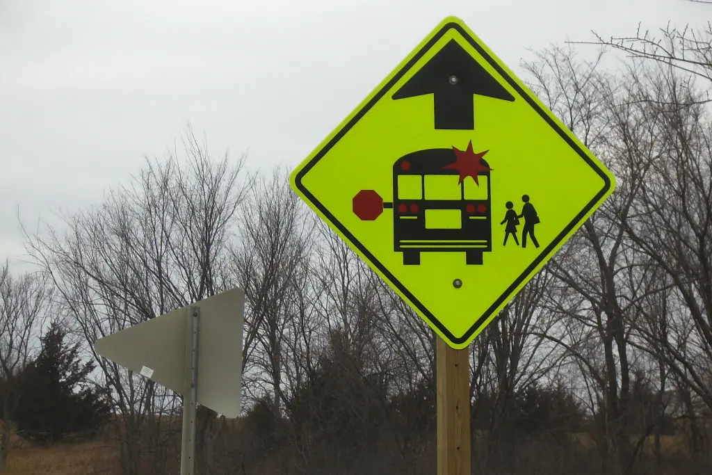 A bus stop caution sign in an unnatural shade of green with trees and a grey sky in the background